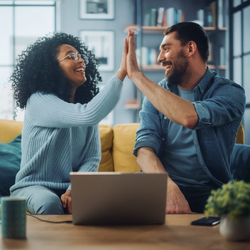 Diverse Multiethnic Couple are Sitting on a Couch Sofa in Stylish Living Room and Choosing Items to Buy Online with Laptop Computer, Give High Five. Friends or Colleagues are Discuss Work Projects. Diverse Multiethnic Couple are Sitting on a Couch Sofa in Stylish Living Room and Choosing Items to Buy Online with Laptop Computer, Give High Five. Friends or Colleagues are Discuss Work Projects.
