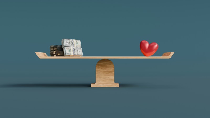 A wooden balance scale holding stacks of hundred-dollar bills on one side and a red heart on the other, illustrating biblical stewardship and the relationship between money and the heart