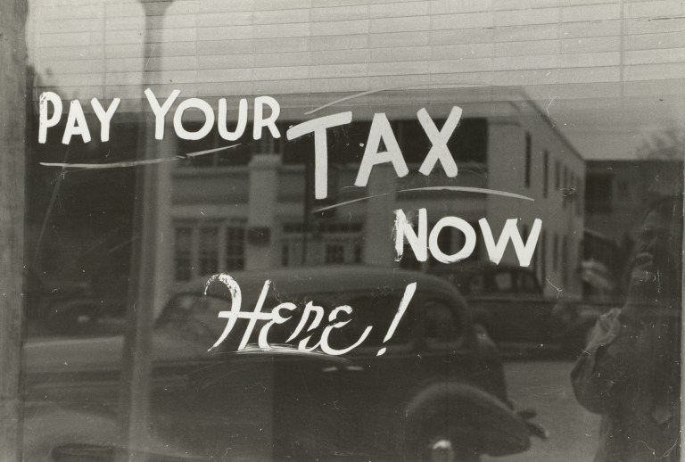 Vintage black-and-white photograph of a storefront window with hand-painted lettering reading "Pay Your Tax Now Here," illustrating an article on the tax benefits of charitable giving for Christian donors.