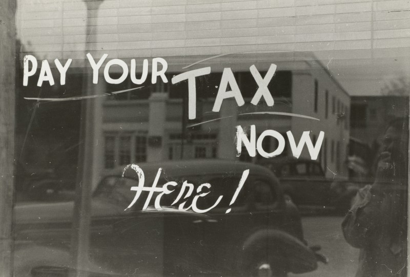 Vintage black-and-white photograph of a storefront window with hand-painted lettering reading "Pay Your Tax Now Here," illustrating an article on the tax benefits of charitable giving for Christian donors.